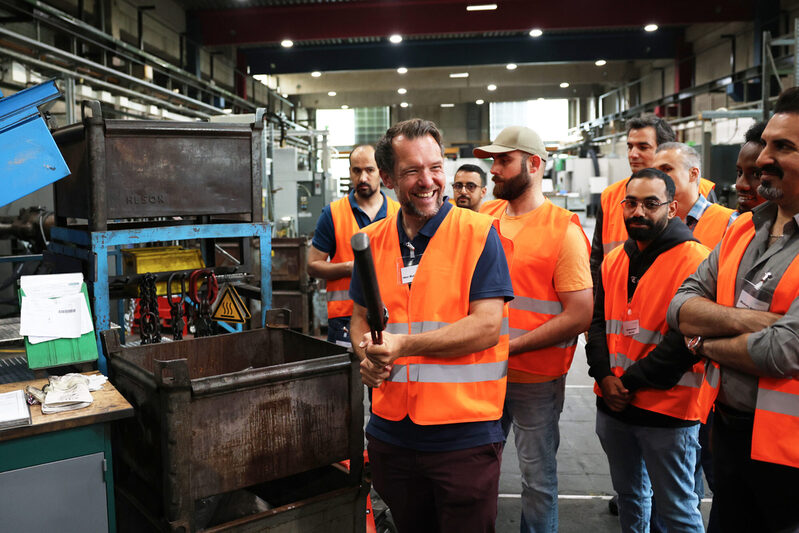 Teilnehmenden mit orangen Sicherheitswesten schauen sich eine Maschine an. Dabei hat der Mitarbeitende der Servicestelle Arbeits- und Fachkräfte ein großes Lachen im Gesicht.