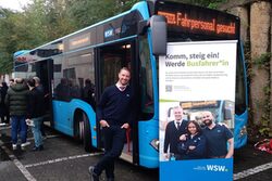 WSW Bus mit Fahrer an der Tür, Gästen im Hintergrund und Roll-Up vor dem Bus mit Werbung zum Busfahrer*in werden